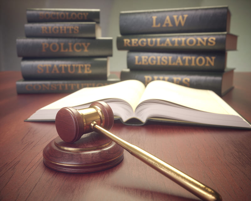 A judge’s gavel lies on a desk beside an open book, with stacks of law, policy, and regulation books in the background.